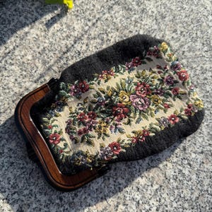 May include: A vintage black purse with a tapestry floral design. The purse features a wooden frame and clasp. The floral pattern includes red, pink, yellow, and green flowers on a cream background.