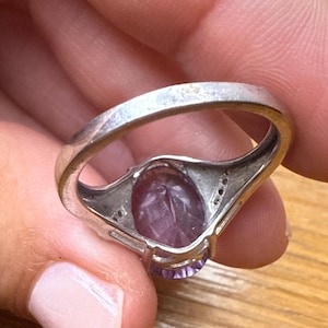 May include: Silver ring with a large oval purple gemstone. The ring has a simple band and small accent stones. The gemstone is a deep purple, and the ring appears to be worn. The ring is likely made of sterling silver.