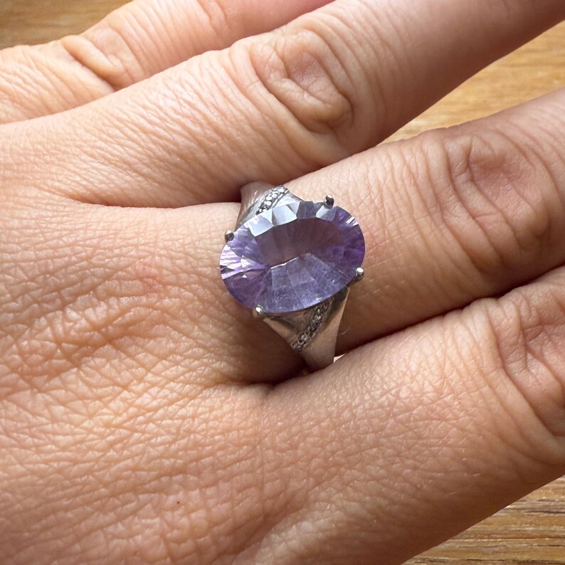 May include: A silver ring with a large, oval-cut, light purple gemstone. The faceted gemstone reflects light. Small, clear stones are set on the ring's band. The ring is worn on a finger.