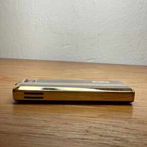May include: A vintage gold and black lighter with horizontal gold stripes. The lighter has a rectangular shape with a gold-colored bottom and a black top. The lighter is resting on a wooden surface.