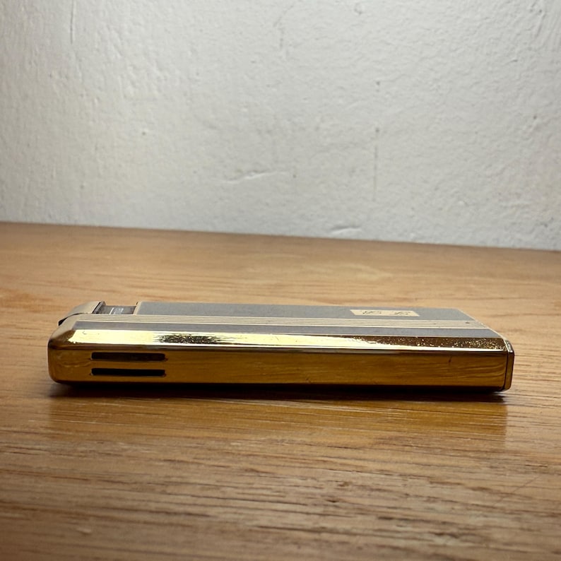 May include: A vintage gold and black lighter with horizontal gold stripes. The lighter has a rectangular shape with a gold-colored bottom and a black top. The lighter is resting on a wooden surface.