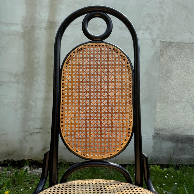 May include: A black wooden chair with a woven cane back and seat. The back of the chair has a distinctive arched design with a circular detail at the top. The woven cane is a light brown color.