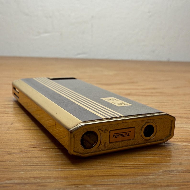 May include: A vintage gold and gray Formula lighter. The lighter has a rectangular shape with a gold frame and a gray top with gold stripes. The word "Formula" is printed on the bottom.