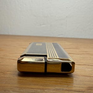 May include: A vintage lighter with a gold-colored metal frame and a black top with gold stripes. The lighter has a rectangular shape and a small opening on one end.