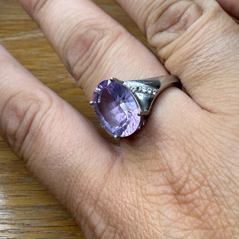 May include: A silver ring with a large, oval-cut, light purple gemstone. The ring has a band with small, clear accent stones. The gemstone is held in place by four prongs.