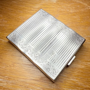 May include: A silver rectangular cigarette case with a textured, striped design on the front and back. The edges feature ornate, swirling patterns. The initials "FB" are visible in a rectangular box on the front.