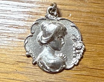 May include: A silver-toned metal pendant featuring a relief profile of a person with an updo hairstyle. The circular pendant has a textured background, a decorative scalloped border, and a small floral cluster on the right side with a top loop.