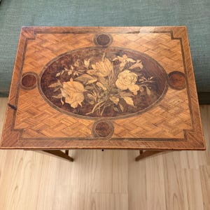 May include: A rectangular wooden table with a floral inlay. The tabletop features a central oval design with roses and other flowers in shades of cream, brown, and green. The surrounding surface has a parquet pattern. The table has four legs.
