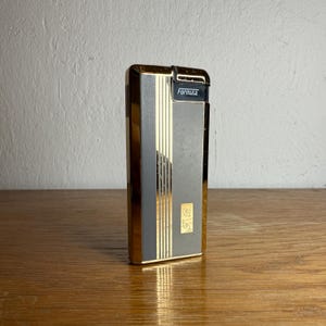 May include: A vintage Formula lighter with a gold-tone frame and a gray and gold striped front. The word "Formula" is printed in black on the top. The lighter has a rectangular shape and is standing on a wooden surface.