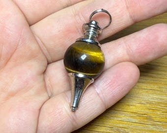May include: A pendulum featuring a polished tiger's eye stone and silver-coloured metal detailing. The pendulum has a rounded, bulbous form with a pointed base and a small ring at the top for hanging.