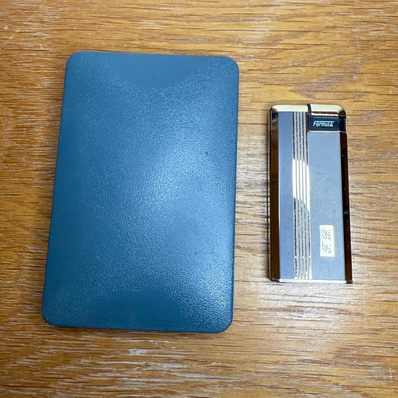 May include: A teal blue rectangular case next to a silver and brown Formula lighter. The lighter has vertical stripes and the text "Formula" on top. The lighter also has the initials "F. B." on the front.