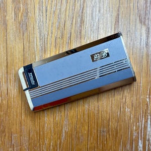 May include: A rectangular, vintage cigarette case with a light blue center and gold-colored trim. The case features horizontal gold stripes and the initials "E.B." in a small gold square. The word "Formula" is printed on the side.