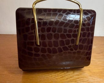May include: A vintage, rectangular handbag with a dark burgundy, faux crocodile skin pattern. It features a gold-coloured, arched handle. The bag is sitting on a wooden surface.