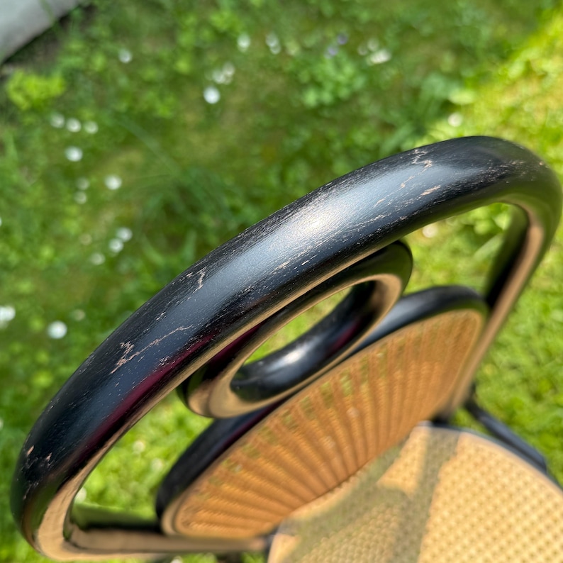 May include: Close-up of a black wooden chair back with a curved design. The chair back has a woven, light brown seat. The chair is set against a blurred green background, suggesting an outdoor setting. The wood shows signs of wear.