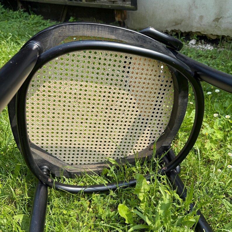 May include: A black wooden chair with a woven cane seat, resting upside down on green grass. The chair's frame is curved, and the seat has a light beige color. The chair is a classic design, suitable for indoor or outdoor use.