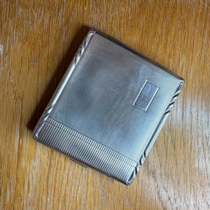 May include: A silver-coloured cigarette case with a textured surface and a rectangular accent. The case has a decorative border and is set against a wooden background. The case is closed.