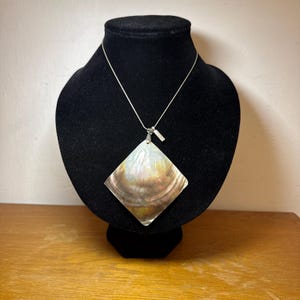 Pode incluir: Um colar de prata com um pingente grande, iridescente e de formato quadrado. O pingente exibe um padrão natural perolado com tons de marrom, branco e dourado. O colar é exibido em um busto de veludo preto.