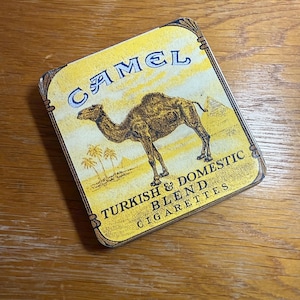 Może przedstawiać: Prostokątna metalowa puszka z żółtym tłem i ilustracją wielbłąda. Słowo "CAMEL" jest w kolorze niebieskim, a tekst "TURKISH & DOMESTIC BLEND CIGARETTES" znajduje się poniżej. Puszka ma wygląd vintage.
