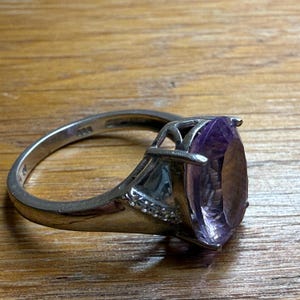 May include: A silver ring with a large, oval-shaped purple gemstone. The ring has a detailed setting with small accent stones and a polished band. The gemstone is the focal point, showcasing its colour and clarity.
