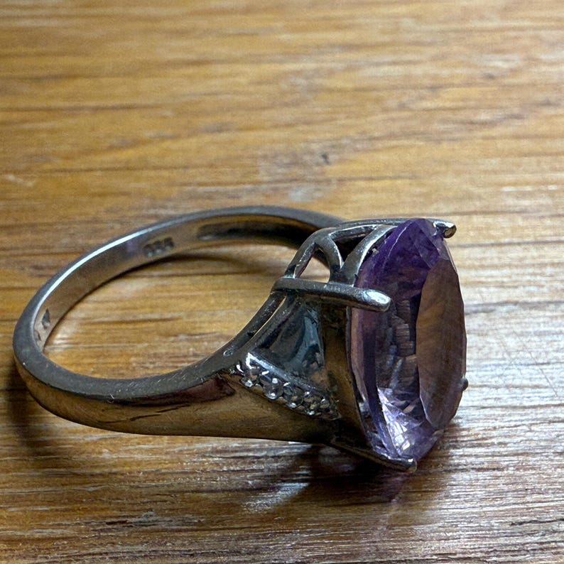 May include: A silver ring with a large, oval-shaped purple gemstone. The ring has a detailed setting with small accent stones and a polished band. The gemstone is the focal point, showcasing its colour and clarity.