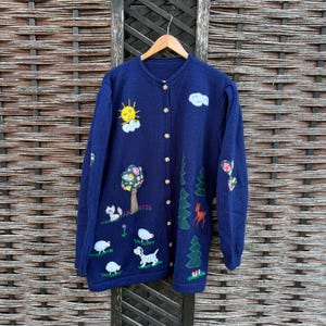 May include: A navy blue cardigan with embroidered countryside motifs. The sweater features a sun, clouds, trees, flowers, sheep, and a dog. It has gold buttons and is displayed on a wooden hanger.
