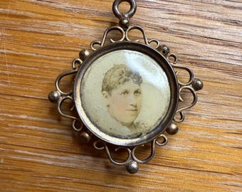 May include: Vintage gold-coloured pendant featuring a portrait of a person. The round portrait is set within a decorative frame with intricate swirls and small ball details. The pendant has a loop at the top for a chain.
