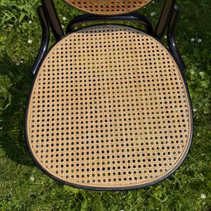 May include: A close-up view of a vintage chair with a woven cane seat and backrest. The chair has a black frame and a curved back. The seat is a light tan color, and the frame is black. The chair is outdoors on a grassy surface.
