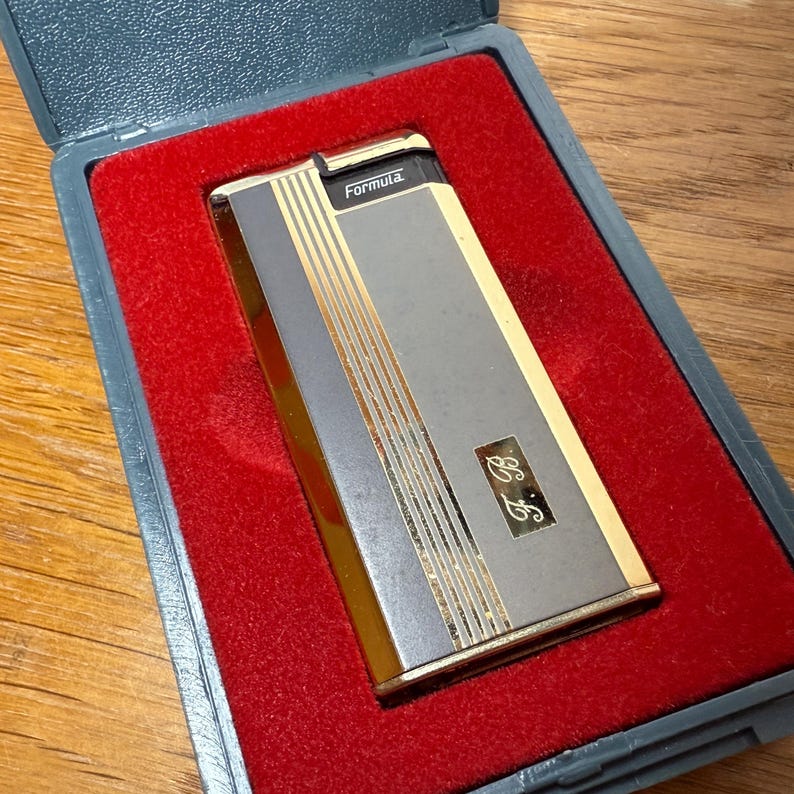 May include: A vintage Formula lighter in a gray case with a red velvet interior. The lighter is silver and gold with vertical gold stripes and the initials "E.F." The word "Formula" is printed on the top.