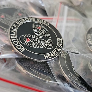 Könnte beinhalten: Eine Sammlung schwarzer und silberner Gedenkmünzen in transparenten Plastiktüten. Die Münzen zeigen den Text "BOOTBLACK EUROPE 2023" und "HEART & SOLE" um ein Schuhabdruck-Design mit einem Herzen.