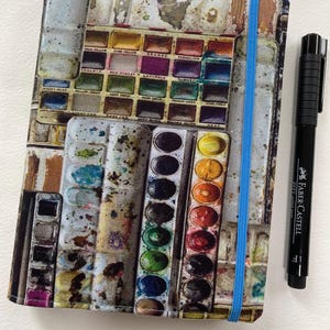 May include: A rectangular notebook featuring a printed design of various watercolour paint sets with pans of different colours and a bright blue elastic closure band, positioned next to a black Faber-Castell Pitt artist pen.