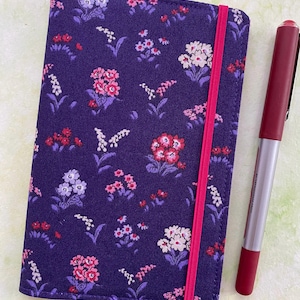 May include: A purple fabric notebook cover with a floral pattern and a pink elastic closure. The cover is next to a red pen with a silver clip.