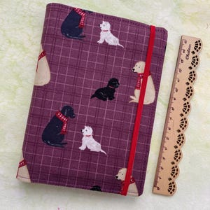 May include: A purple fabric notebook cover with a red elastic closure. The cover features a repeating pattern of dogs wearing scarves, in black, white, and brown.