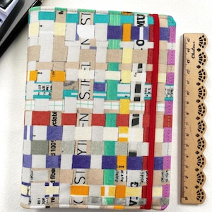 May include: A rectangular notebook with a woven cover made of multicoloured paper strips featuring printed text. The notebook is secured by a vertical red elastic band and is positioned next to a wooden ruler measuring 15 centimetres in length.