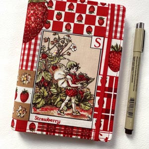May include: A notebook with a red and white gingham pattern and a strawberry design. The cover includes a vintage illustration of a child gathering strawberries, with the word "Strawberry" below. A red elastic band secures the notebook. A pen is placed next to it.