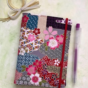 May include: A patterned fabric book cover with a patchwork design. The cover features floral and geometric patterns in pink, red, blue, and gold. A red elastic band secures the cover. A purple pen is placed beside it.