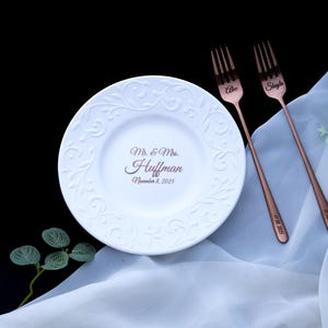 May include: A white ceramic plate with an embossed scroll pattern and the text Mr. & Mrs. Huffman November 8, 2025, paired with two copper-colored metal forks engraved with the names Alex and Shayla and the date 11.08.2025.
