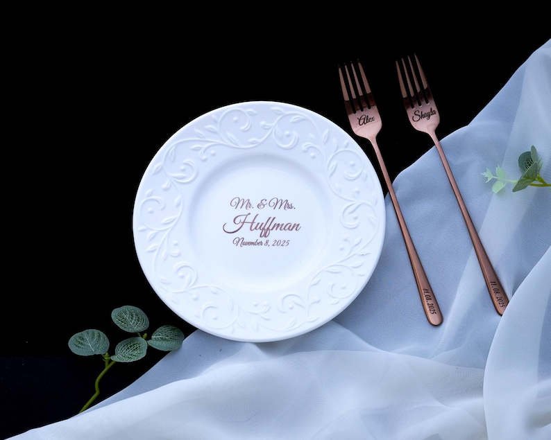 May include: A white ceramic plate with an embossed scroll pattern and the text Mr. & Mrs. Huffman November 8, 2025, paired with two copper-colored metal forks engraved with the names Alex and Shayla and the date 11.08.2025.
