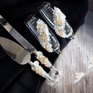 May include: A wedding cake cutting knife and two champagne flutes decorated with seashells and beads. The knife is engraved with "Mr. & Mrs. Lowe Jr." and the flutes are engraved with "Love Is... 01.2023" and "Love Is... 2023".