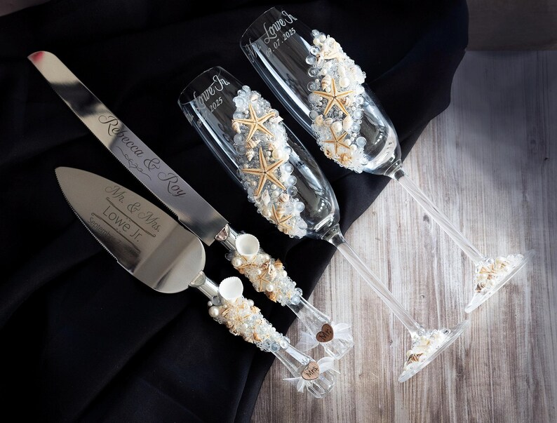May include: A wedding cake cutting knife and two champagne flutes decorated with seashells and beads. The knife is engraved with "Mr. & Mrs. Lowe Jr." and the flutes are engraved with "Love Is... 01.2023" and "Love Is... 2023".