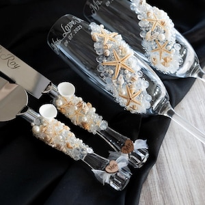 May include: A wedding cake cutting set with a silver knife and server, decorated with seashells and pearls. The champagne flutes are also decorated with seashells and pearls. The knife and server are engraved with the names "Rebecca & Roy" and "Mr. & Mrs. Lowe Jr." and the date "September 7, 2023".