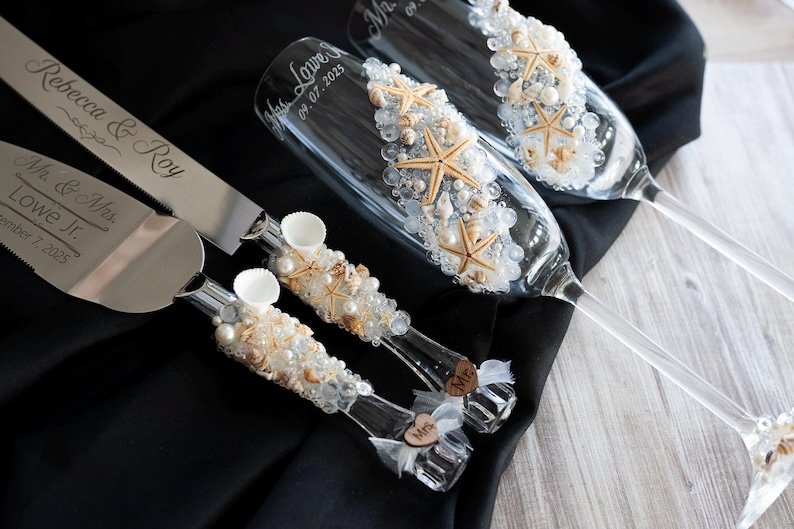 May include: A wedding cake cutting set with a silver knife and server, decorated with seashells and pearls. The champagne flutes are also decorated with seashells and pearls. The knife and server are engraved with the names "Rebecca & Roy" and "Mr. & Mrs. Lowe Jr." and the date "September 7, 2023".