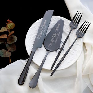 May include: A set of matte black stainless steel cake serving utensils including a knife, a cake server, and two forks. The knife is engraved with "Melanie & Cameron 10.04.2025", the server with "Mr. & Mrs. Skinner October 4, 2025", and the forks with names.