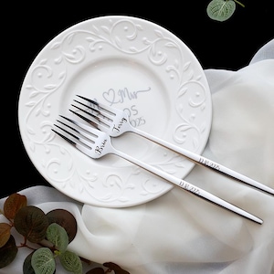 May include: Two silver-toned metal forks with engraved names Bria and Jerry on the tines and the date 08.16.2025 on the handles, resting on a white ceramic plate with an embossed scroll pattern and the text Mr & Mrs 08.16.2025.