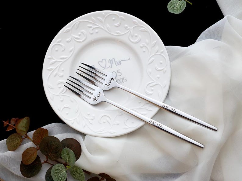 May include: Two silver-toned metal forks with engraved names Bria and Jerry on the tines and the date 08.16.2025 on the handles, resting on a white ceramic plate with an embossed scroll pattern and the text Mr & Mrs 08.16.2025.