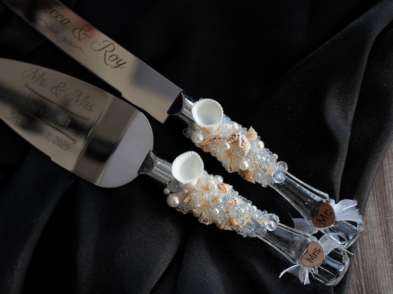 May include: A set of two silver cake knives with white and gold shell and pearl embellishments. The handles are decorated with the words "Mr." and "Mrs." and the blade has the text "Erica & Roy" engraved on it.
