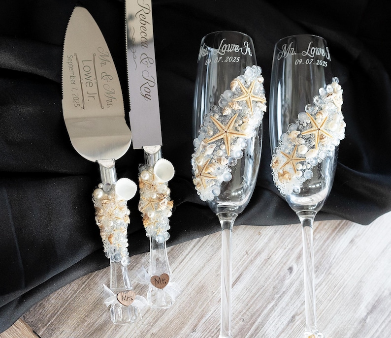 May include: A wedding cake cutting set with a silver knife and two champagne flutes decorated with seashells and starfish. The knife has the text "Mr. & Mrs. Lowe Jr. September 7, 2025" engraved on the blade. The flutes have the text "Mr. Lowe Jr. 07.07.2025" and "Mr. Lowe 09.07.2025" engraved on them.