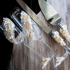 May include: Two champagne flutes decorated with seashells and starfish, a silver cake knife and server with matching seashell and starfish handles, and a small wooden sign with the text "Mr. & Mrs. Lowe Jr. September 7, 2025".