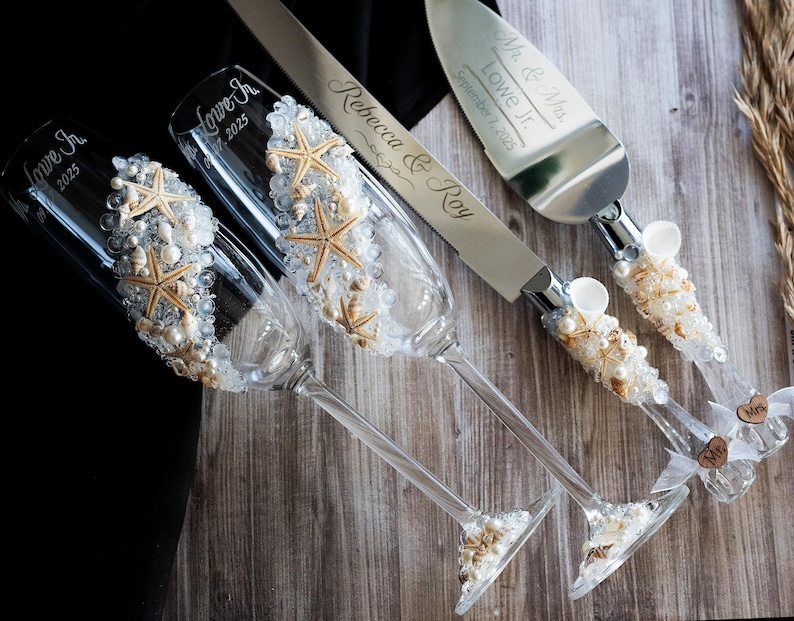 May include: Two champagne flutes decorated with seashells and starfish, a silver cake knife and server with matching seashell and starfish handles, and a small wooden sign with the text "Mr. & Mrs. Lowe Jr. September 7, 2025".