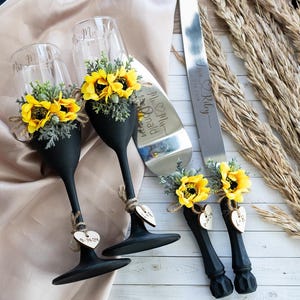 May include: Black painted champagne flutes with sunflower accents and wooden heart charms with the date "06.04.2024" engraved on them. A black painted cake knife with a sunflower accent and a wooden heart charm with the text "Mr. & Mrs. Riley" engraved on it.