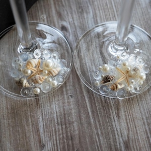 May include: Two clear glass wine glasses with white pearl beads and seashells inside. The glasses are on a wooden surface.